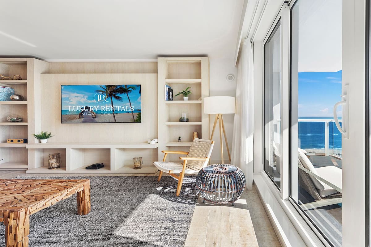 A modern living room with a large TV, wooden shelves, a floor lamp, and a view of the ocean.