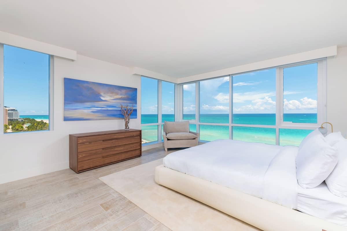 A bedroom with a large bed, a dresser, a chair, and a painting. It has large windows with a view of the ocean.