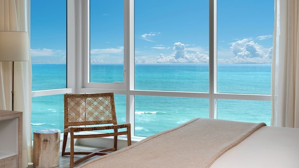 Ein Zimmer mit großem Fenster zum Meer, ein Korbstuhl und ein Bett mit beiger Decke.