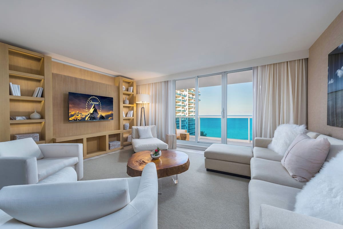 A modern hotel room with a large balcony, a flat-screen TV, a wooden coffee table, and a bookshelf.