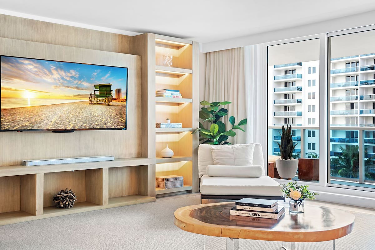 A modern living room with a large flat-screen TV mounted on a wooden wall, a white sofa, a round coffee table with books, and a large window offering a view of a high-rise building.