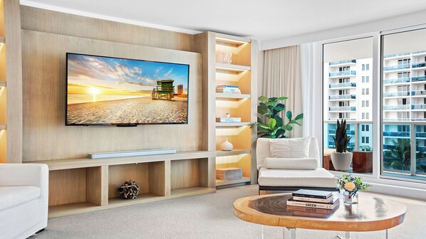 A modern living room with a flat-screen TV, a white sofa, a coffee table, and a bookshelf. - Luxury Vacation Rentals at 1 Hotel & Homes by LRMB (Miami Beach)