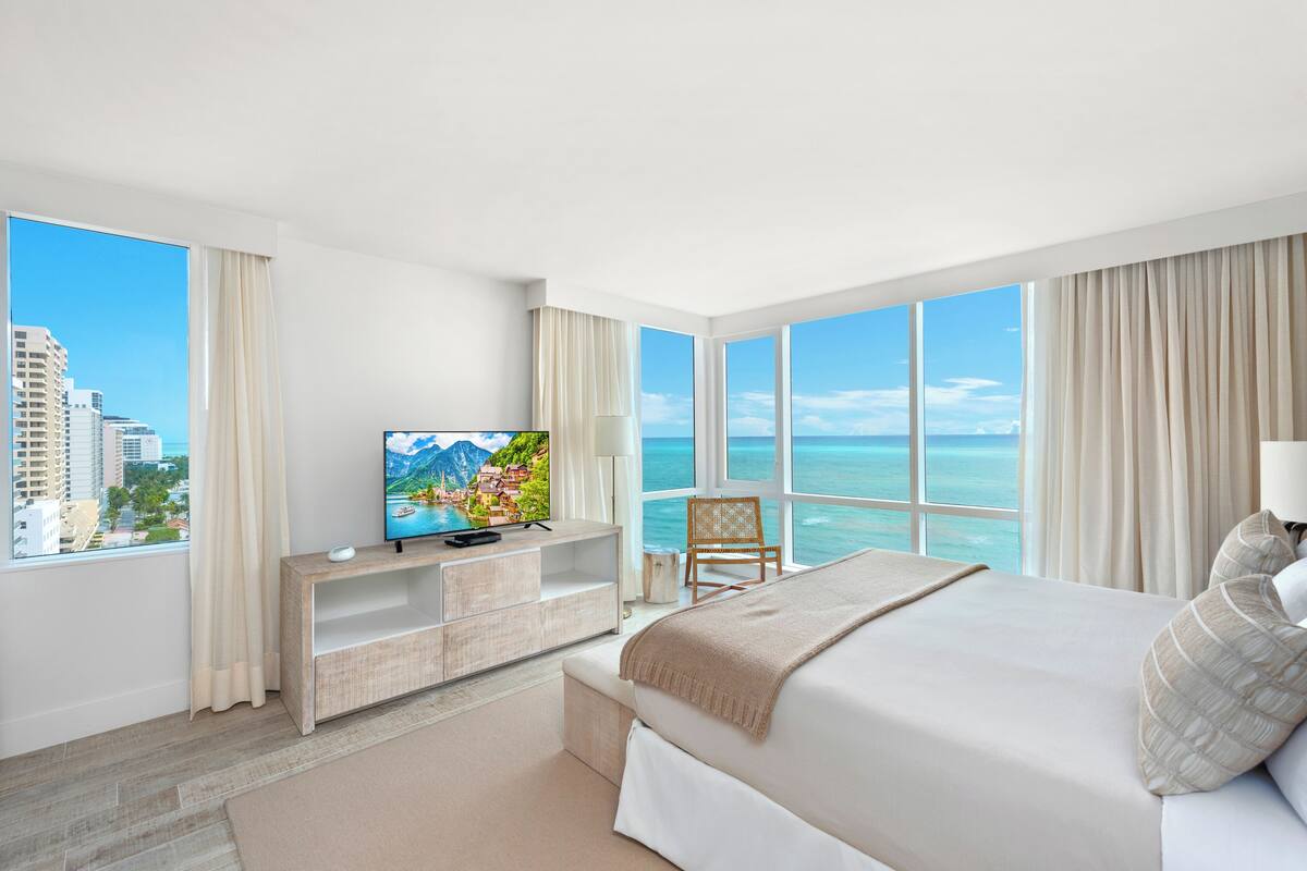 A hotel room with a large bed, a TV on a wooden console, a chair, and a view of the ocean.