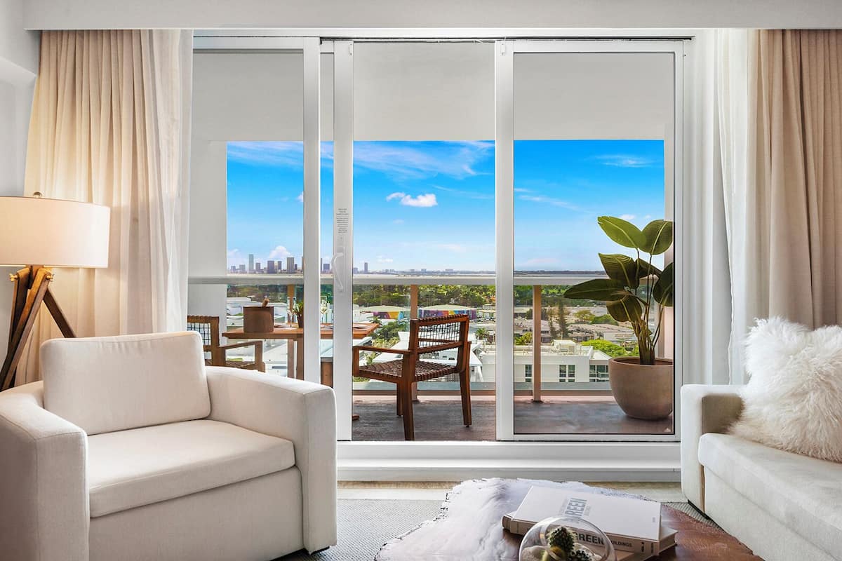 A modern living room with a sofa, a chair, a lamp, and a view of a cityscape through large glass doors.