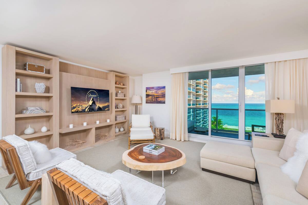 A modern living room with a large TV, comfortable seating, a wooden coffee table, and a balcony with a view of the ocean.