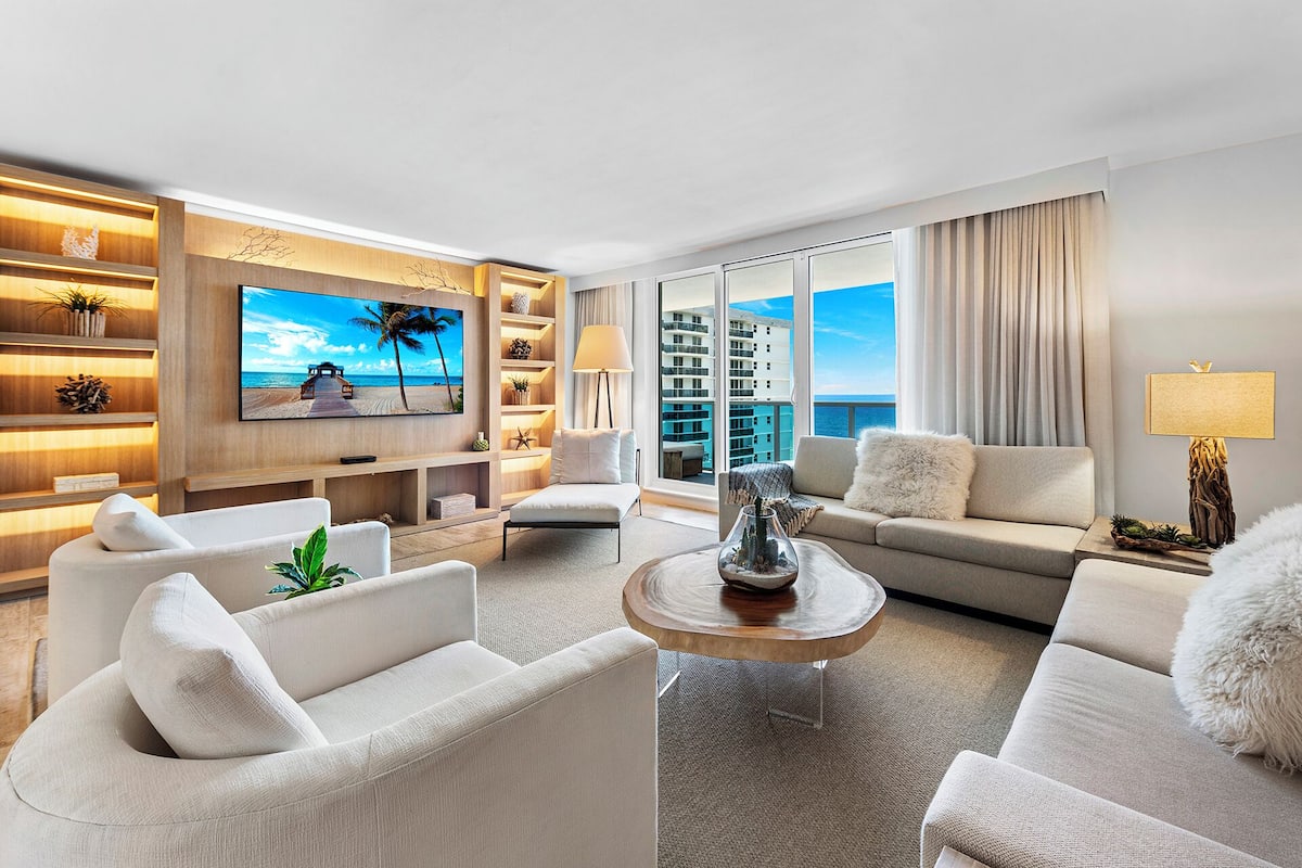 A modern living room with a flat-screen TV, comfortable seating, a coffee table, and a view of the ocean through large windows.