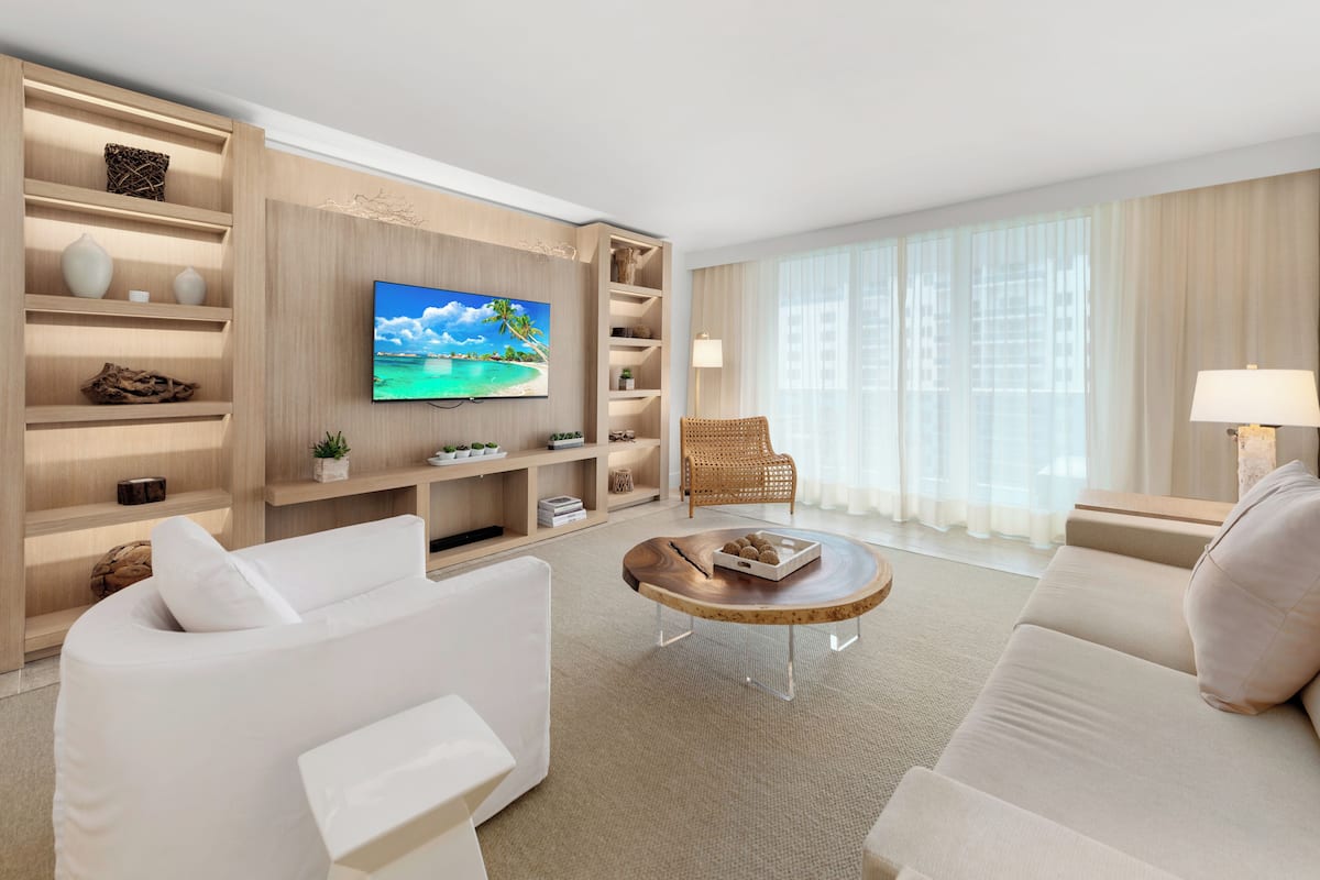 A modern living room with a flat-screen TV, white sofas, a wooden coffee table, and a wicker chair.