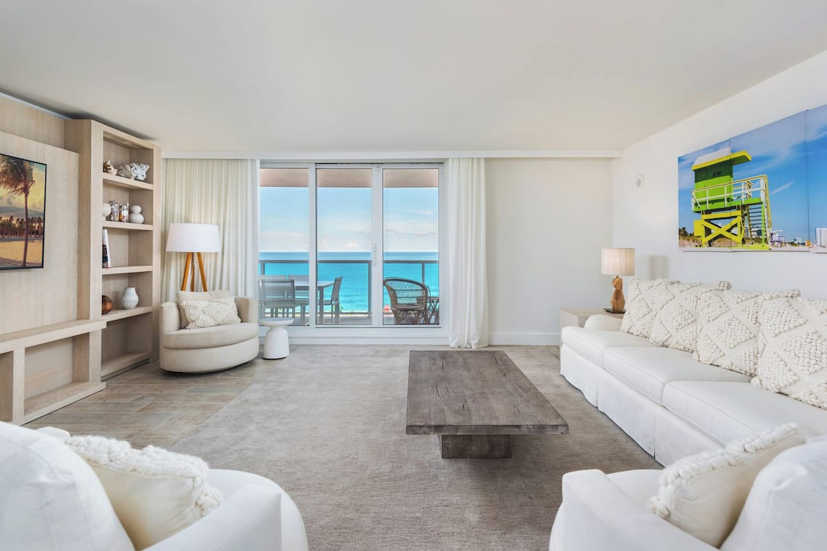 A spacious living room with a large sliding glass door leading to a balcony with a view of the ocean. There is a white sofa, a coffee table, and a bookshelf with decorative items.