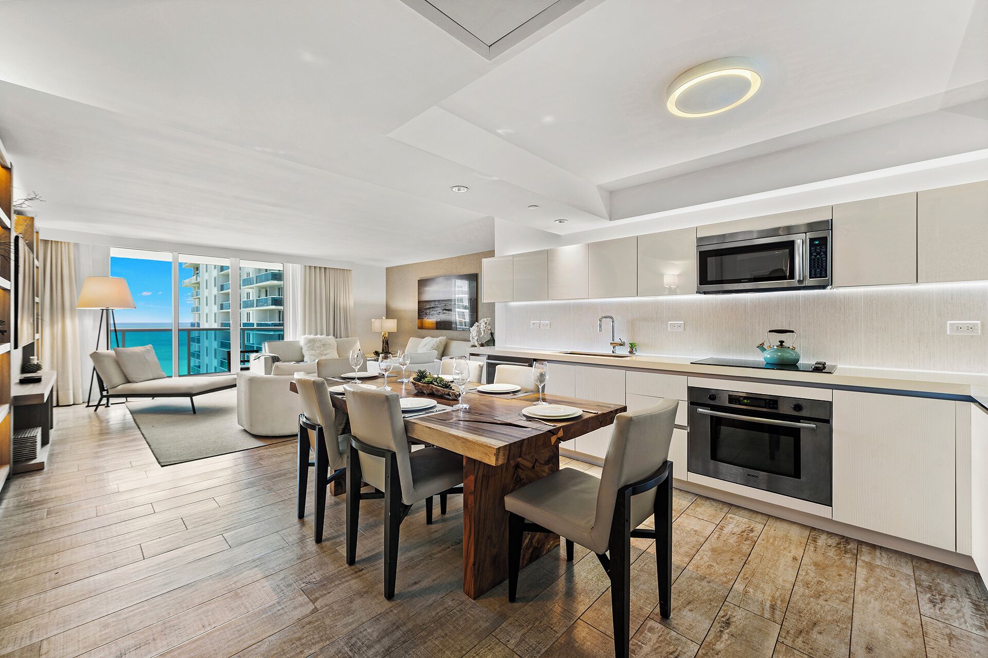 A modern hotel room with a dining area, kitchen, and a balcony with a view of the sea.