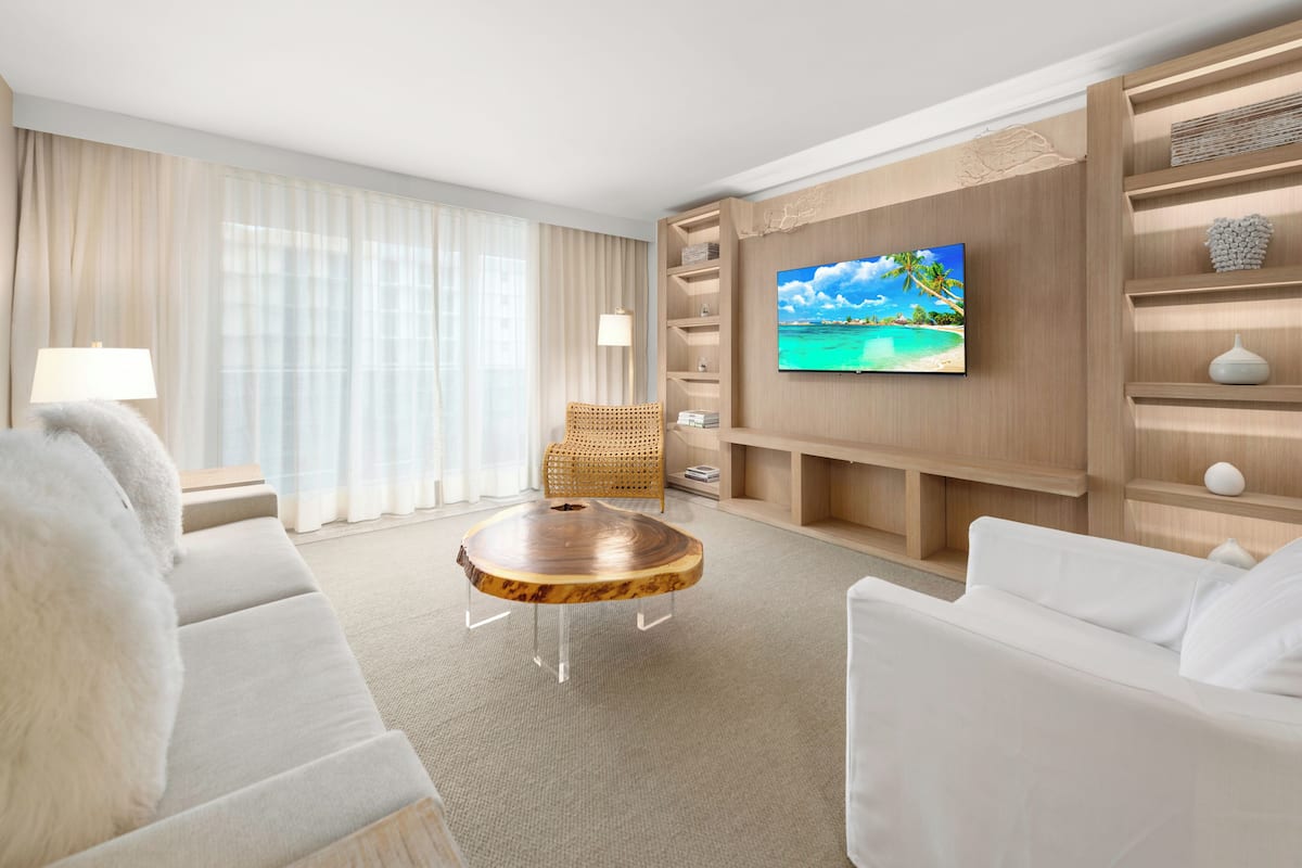 A modern hotel room with a large flat-screen TV mounted on the wall, a comfortable sofa, a round coffee table, and a wicker chair.