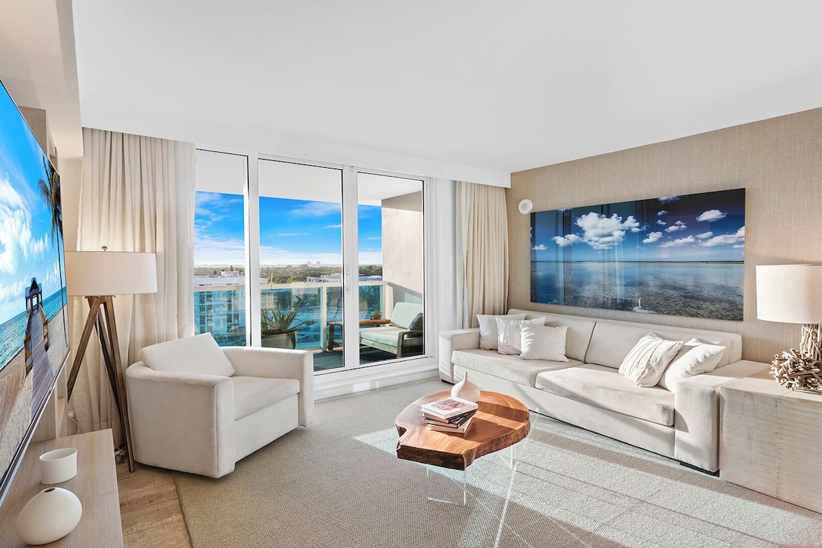 A modern hotel room with a large balcony, white furniture, and a large painting of a beach scene.