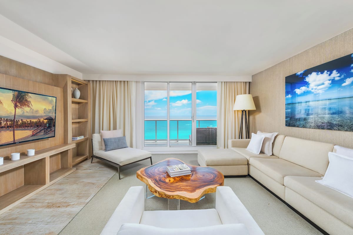 A modern hotel room with a large window offering a view of the ocean, a wooden coffee table, a sofa, a chair, and a TV on the wall.