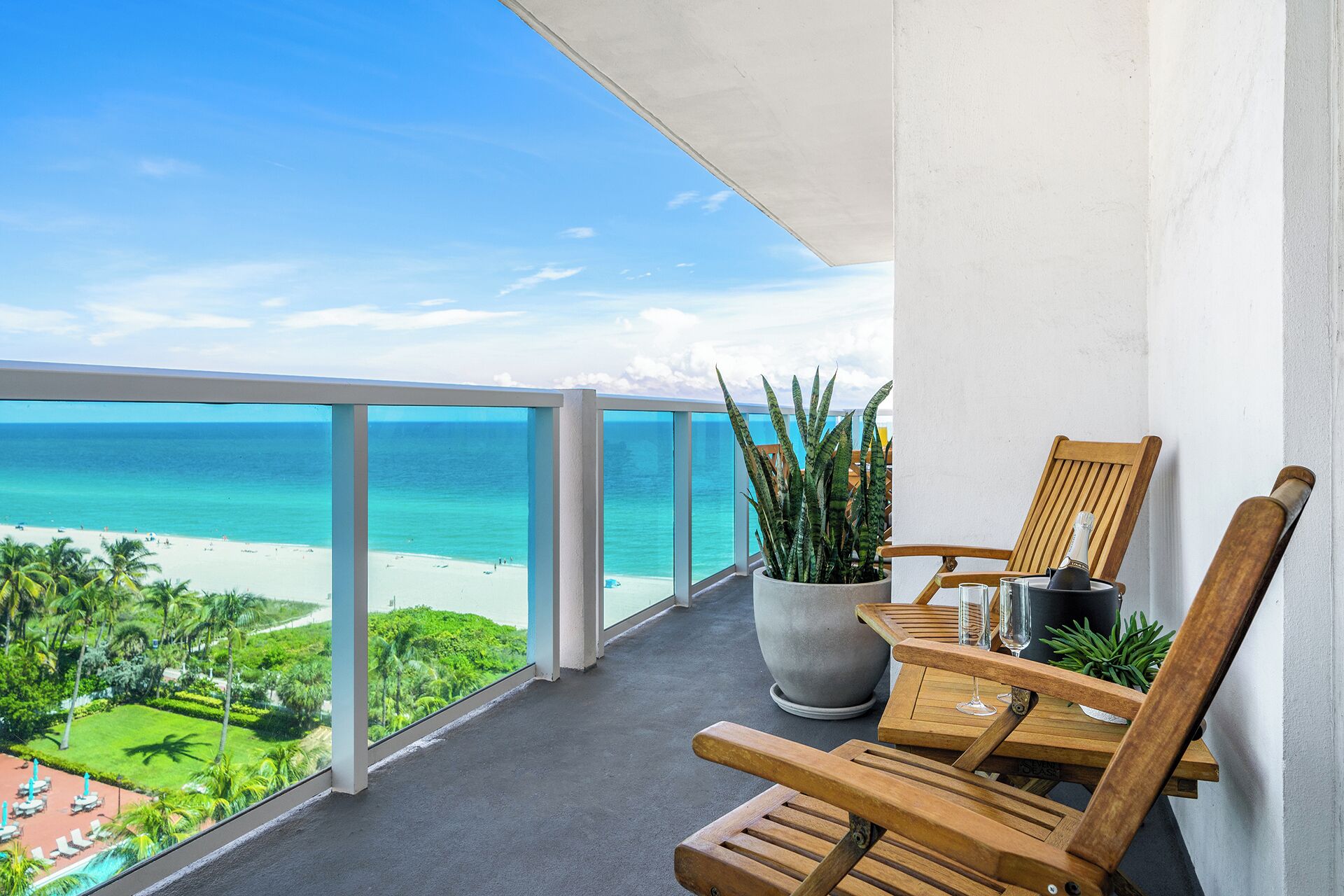 A balcony with a view of the beach, a wooden chair, a potted plant, and a bottle of champagne.