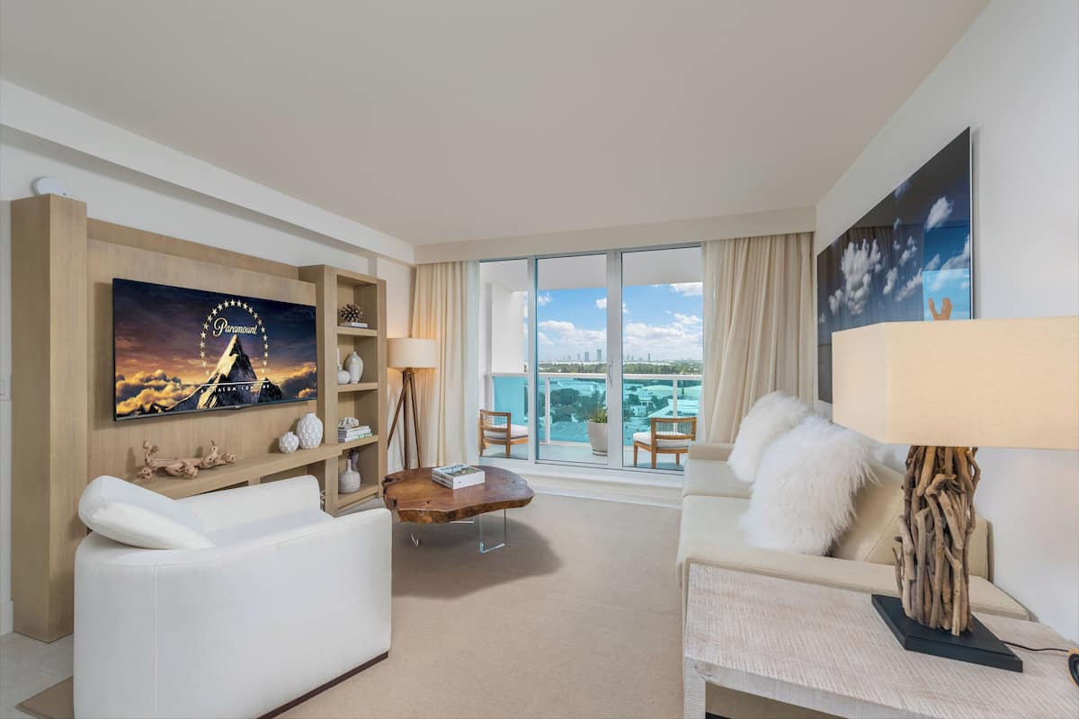 A modern hotel room with a large window offering a city view, a flat-screen TV mounted on the wall, and a cozy seating area with a sofa and armchair.