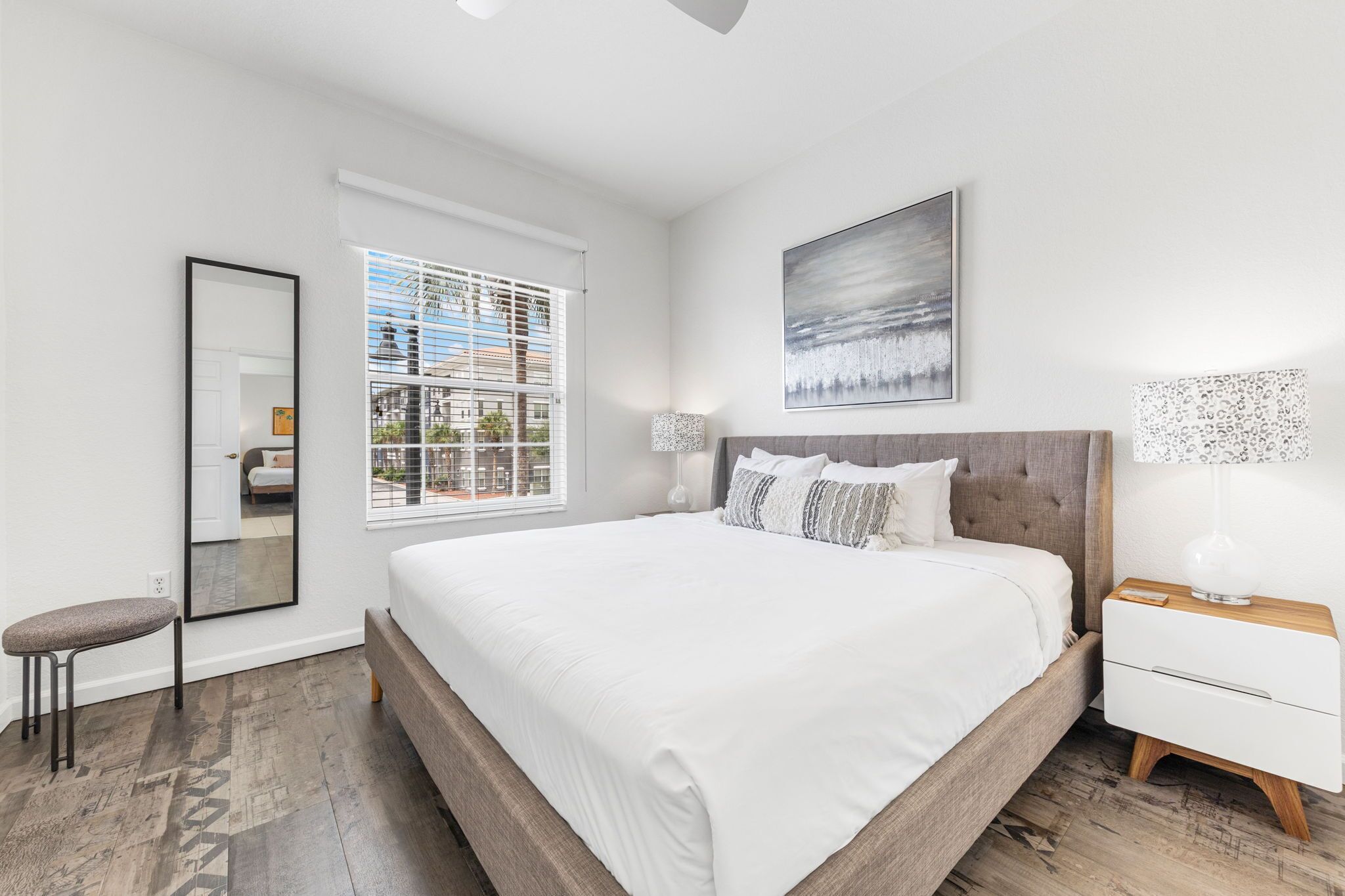 A modern bedroom with a large bed, a bedside table, a nightstand, a mirror, and a window with blinds.