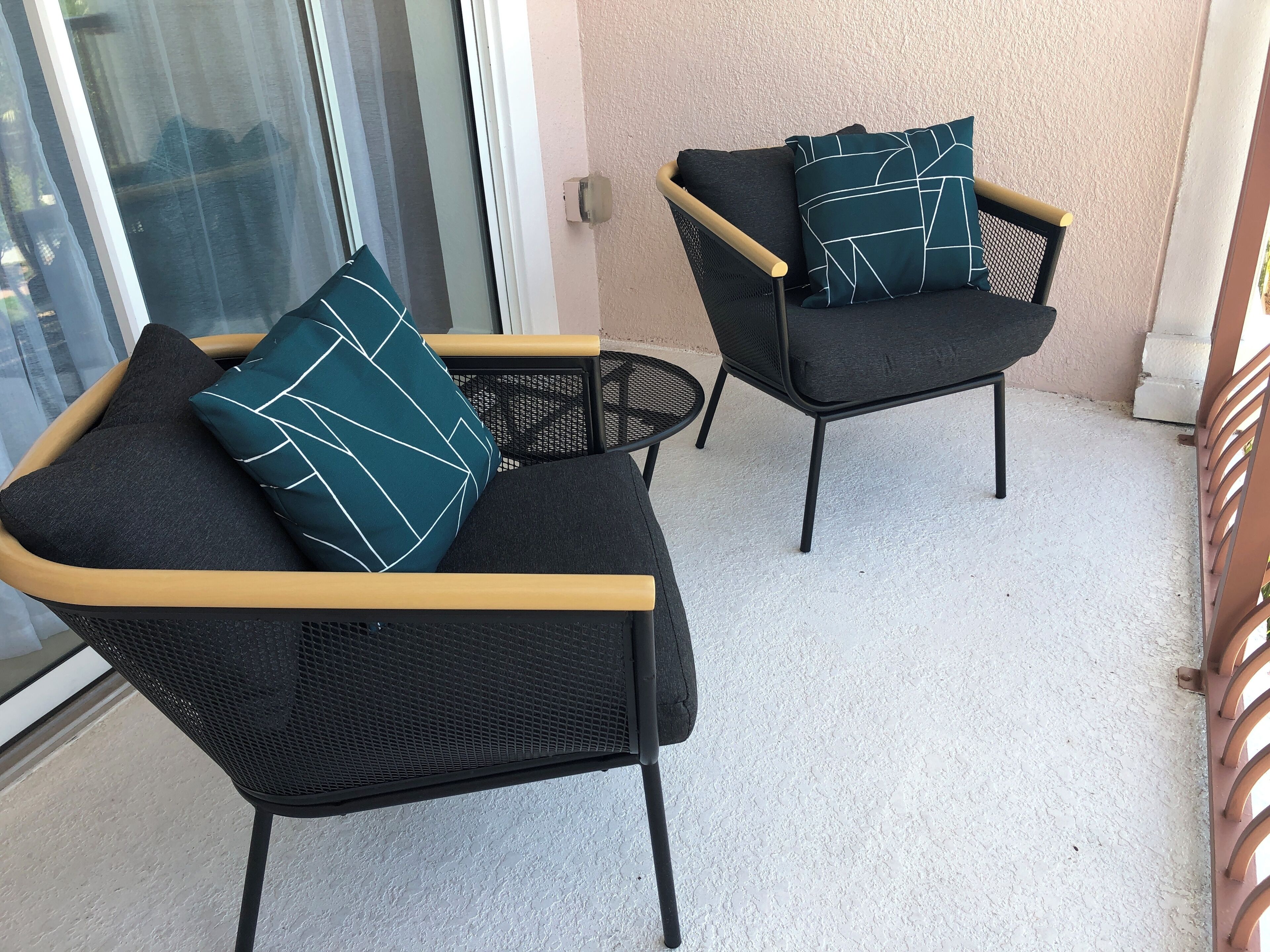Two black wicker chairs with wooden armrests and teal pillows, placed on a balcony with a textured wall and a glass door in the background.