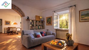 A cozy living room with a striped sofa, a wooden coffee table with fruit, a bookshelf, and a window with sunlight. - Il Palazzaccio | Nature, Pool & Relaxation in Lucca (Lucca)