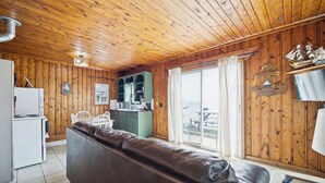 Ein gemütliches Blockhaushaus-Interieur mit Holzbalkendecke, Ledersofa, Küchenzeile und großem Fenster mit Vorhängen.