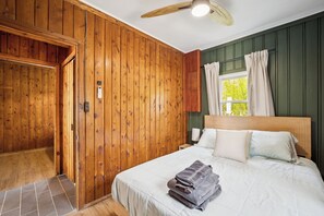 A bedroom with wooden walls, a bed with white bedding, a nightstand, a lamp, and a window with curtains.