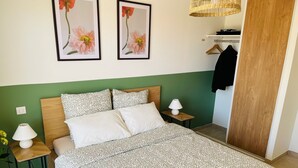 Une chambre bien rangée, avec un lit, deux oreillers, une table de chevet avec une lampe, une petite table sur laquelle est posé un vase de fleurs, et deux tableaux représentant des motifs floraux, accrochés au mur.