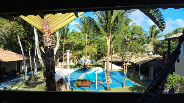 A pool area with a hammock, palm trees, and thatched-roof buildings. - Pousada Água Marinha (Porto Seguro)