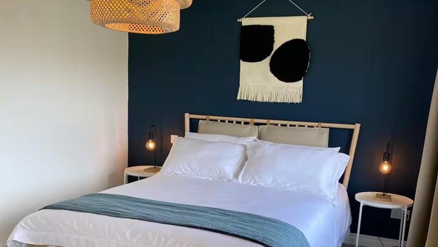 Una cama con sábanas blancas, cabecera de madera y un banco al pie. Hay dos mesitas de noche con lámparas.