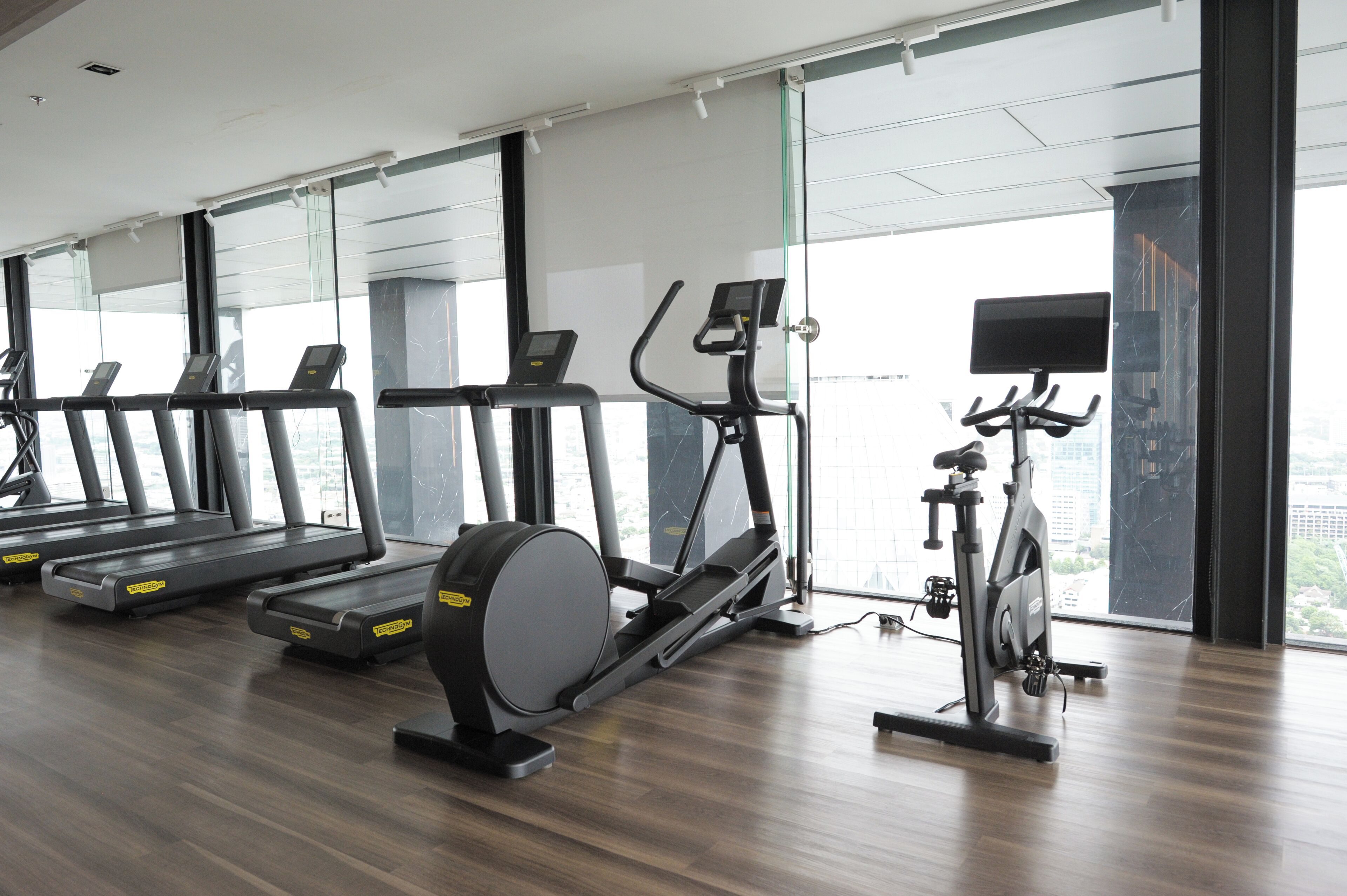 A gym with treadmills, an elliptical, and a stationary bike, large windows, and a city view.