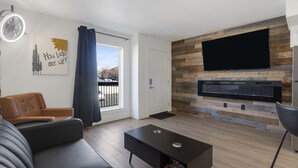 Ein modernes Wohnzimmer mit einem Flachbildfernseher an einer Holzwand, einem Ledersofa, einem Couchtisch und einem großen Fenster mit Vorhängen.
