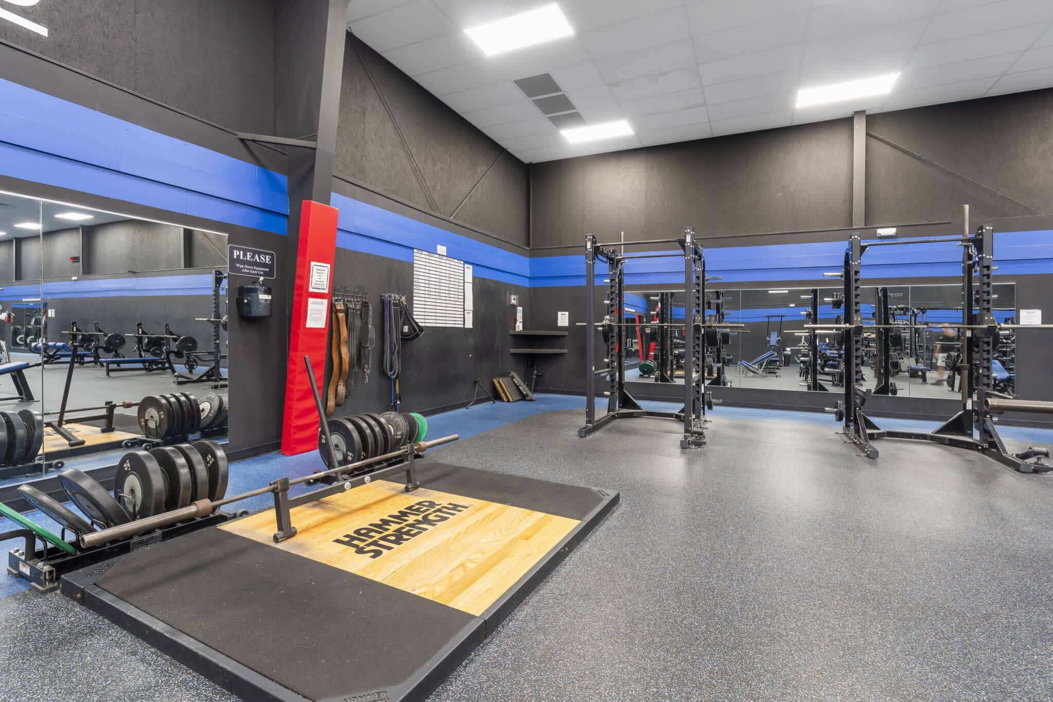 Un gimnasio con equipamiento de levantamiento de pesas, espejos y una plataforma de madera con el texto 'FIRMER STRENGTH'.