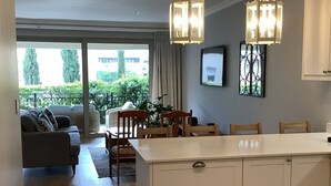 A modern kitchen with white cabinets, a central island, and pendant lighting. There is a dining area with a table and chairs, and a living room with a sofa and a large window overlooking a garden.