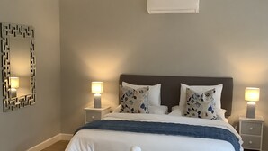 A neatly arranged hotel room with a bed, bedside lamps, a bedside table, a wall-mounted air conditioner, and a decorative mirror.