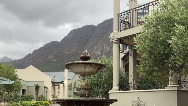 A stone fountain with water cascading into a basin, surrounded by well-maintained greenery and a paved path leading to a building with a balcony.
