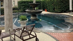 A pool with a stone fountain, surrounded by lounge chairs and a potted plant.