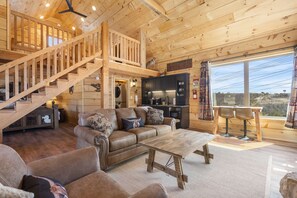 A wooden cabin interior with a staircase, a living room with a sofa and coffee table, a kitchen area, and a large window with a view. - Elk Lodge Cabin for 4! (Branson)