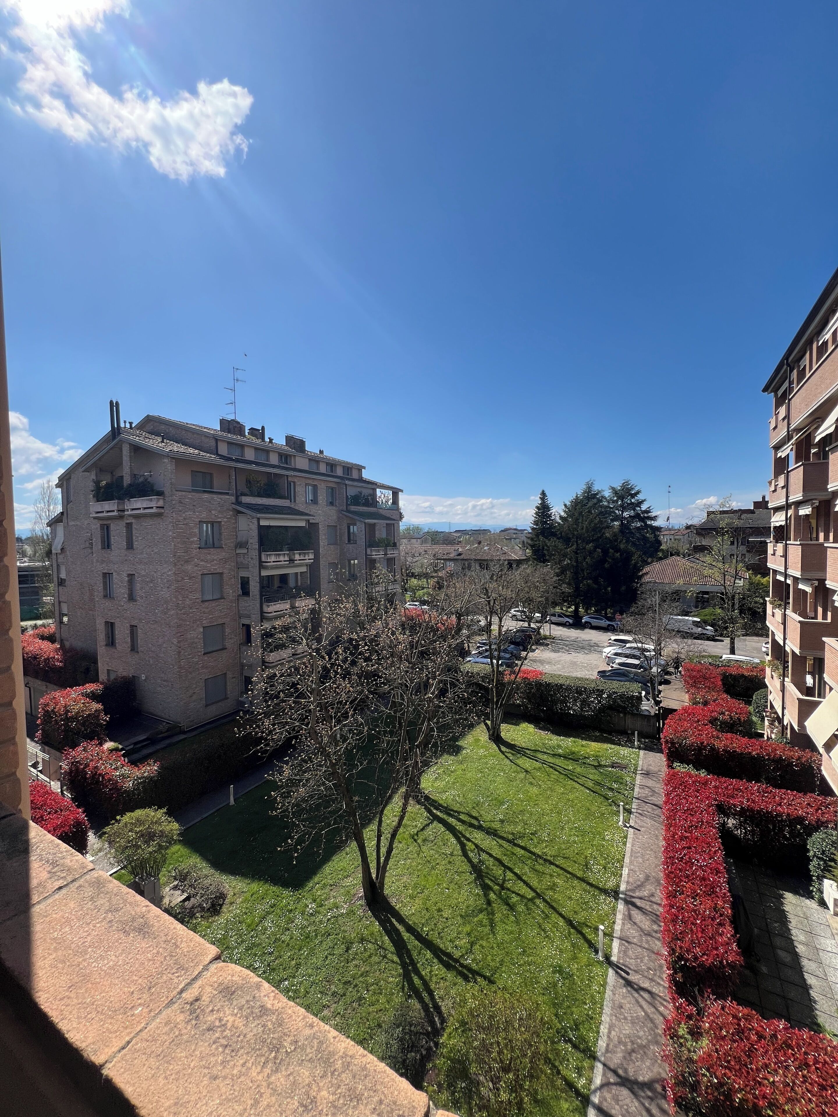 A residential building with a stone facade, multiple balconies, and a well-maintained garden with red flowering bushes.