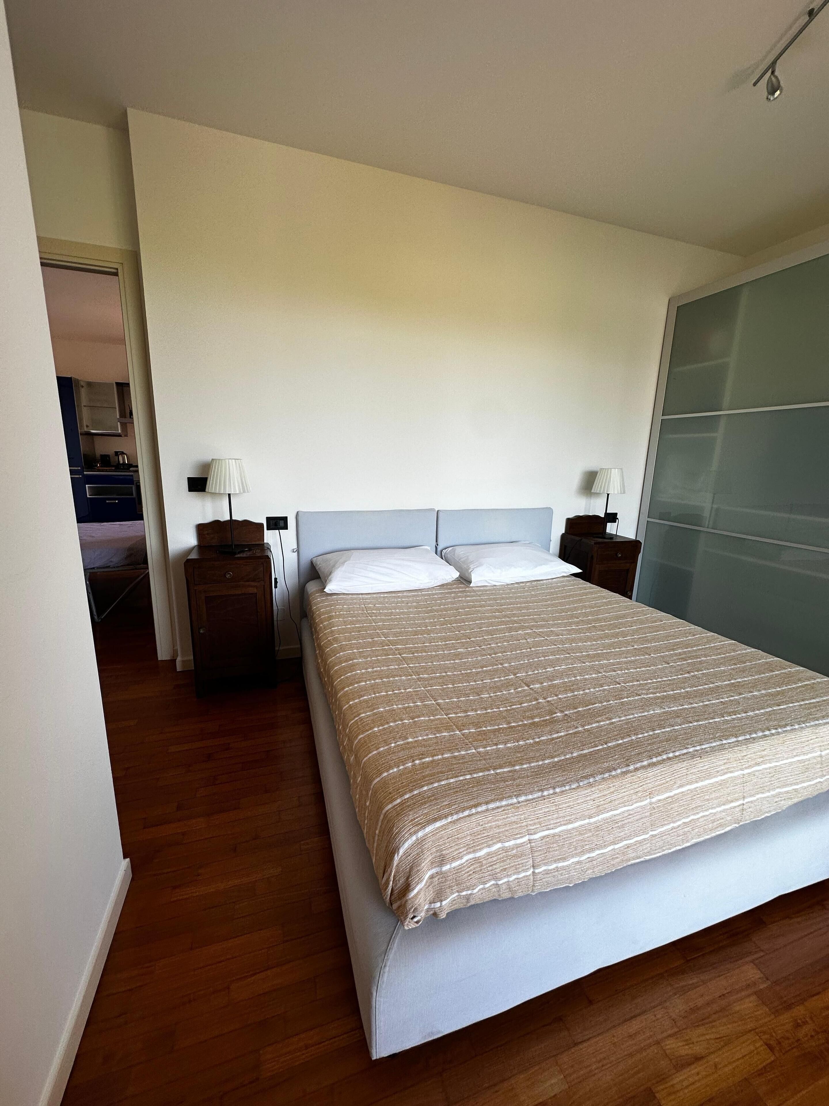 A neatly arranged bedroom with a large bed, two bedside tables, and a view of a kitchen area.