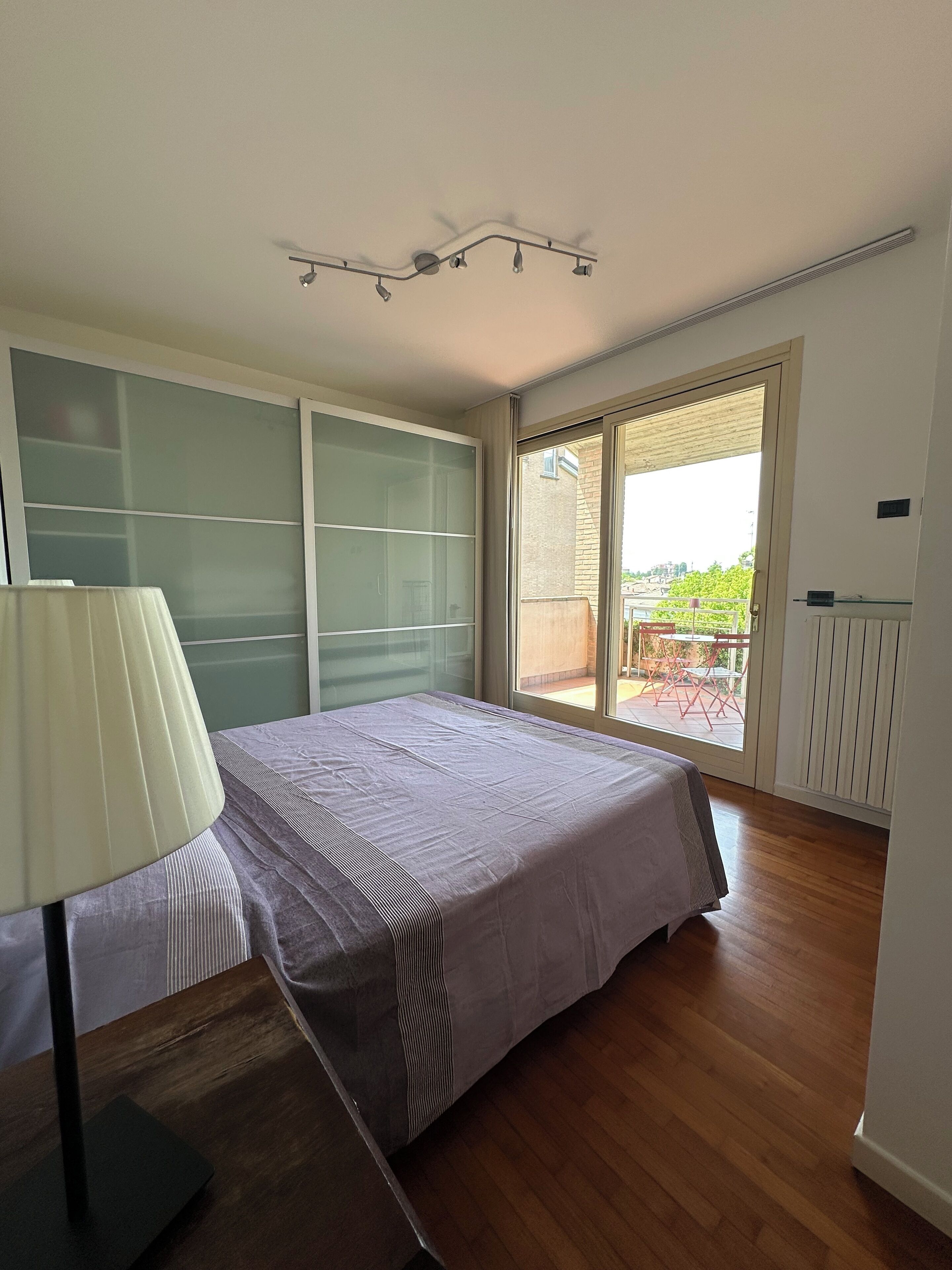 Ein Schlafzimmer mit einem Bett, einem Nachttisch, einer Lampe, einer Schiebetür zu einem Außenbereich und einer Deckenleuchte.