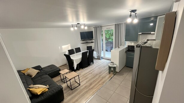 A modern living room with a black sofa, a dining table with chairs, a small kitchen area, and a TV mounted on the wall. - Bright and Spacious 3-Bedroom Apartment - Direct Access to Central Paris (Choisy-le-Roi)