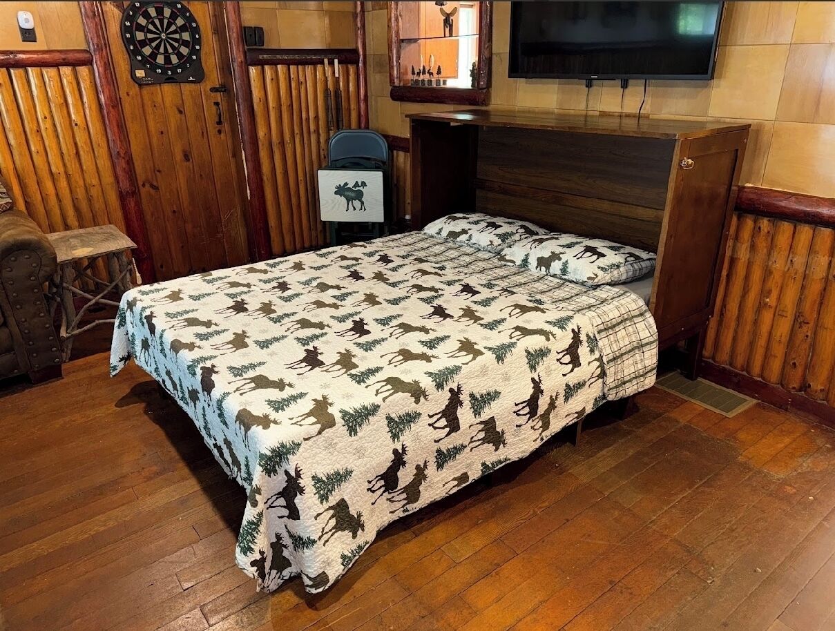 Une chambre confortable avec un lit agrémenté d’une couvre-lit à motifs d’élans, un jeu de fléchettes et un téléviseur fixé au mur.