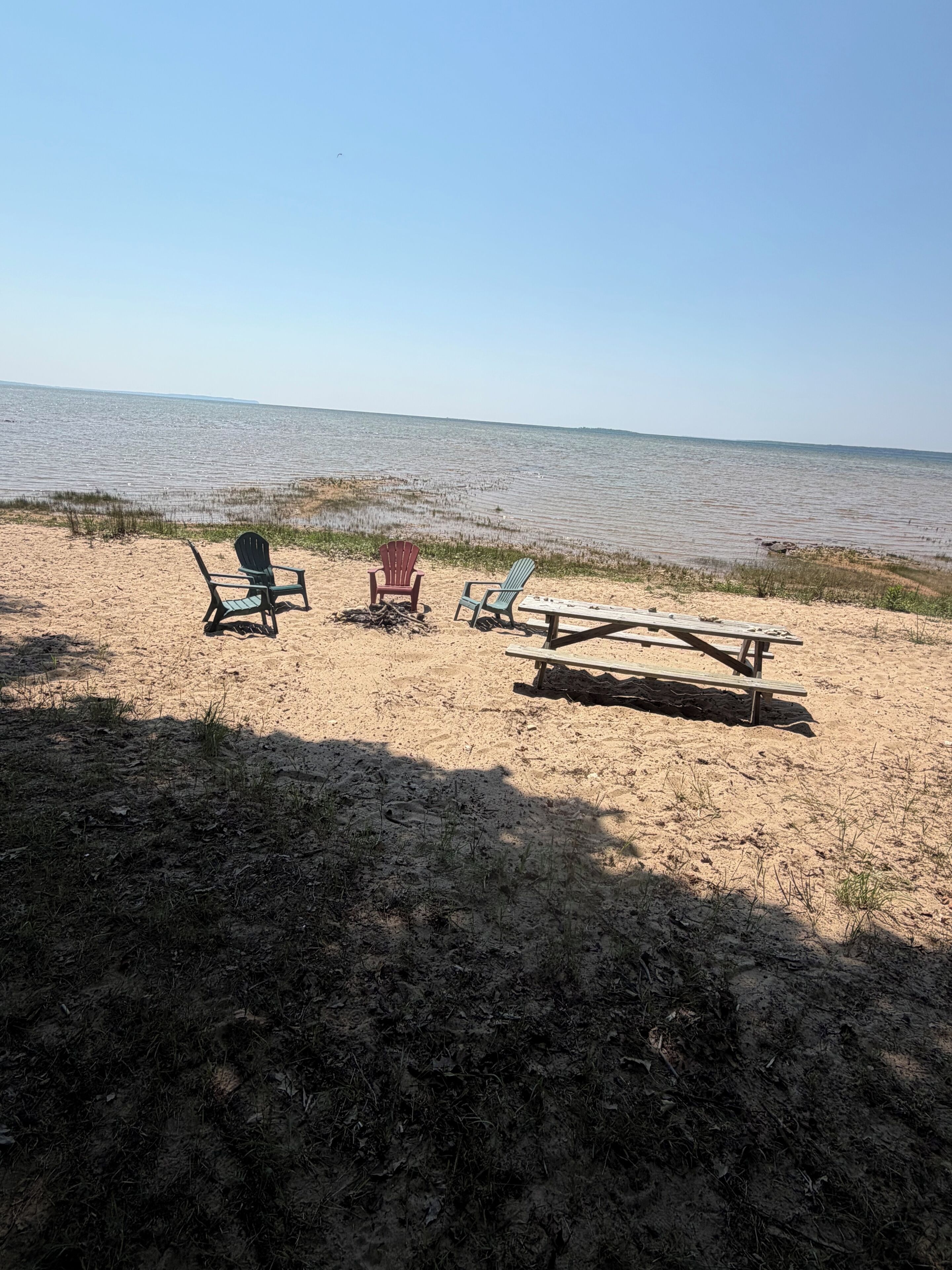 A beach with three Adirondack chairs and a picnic table.