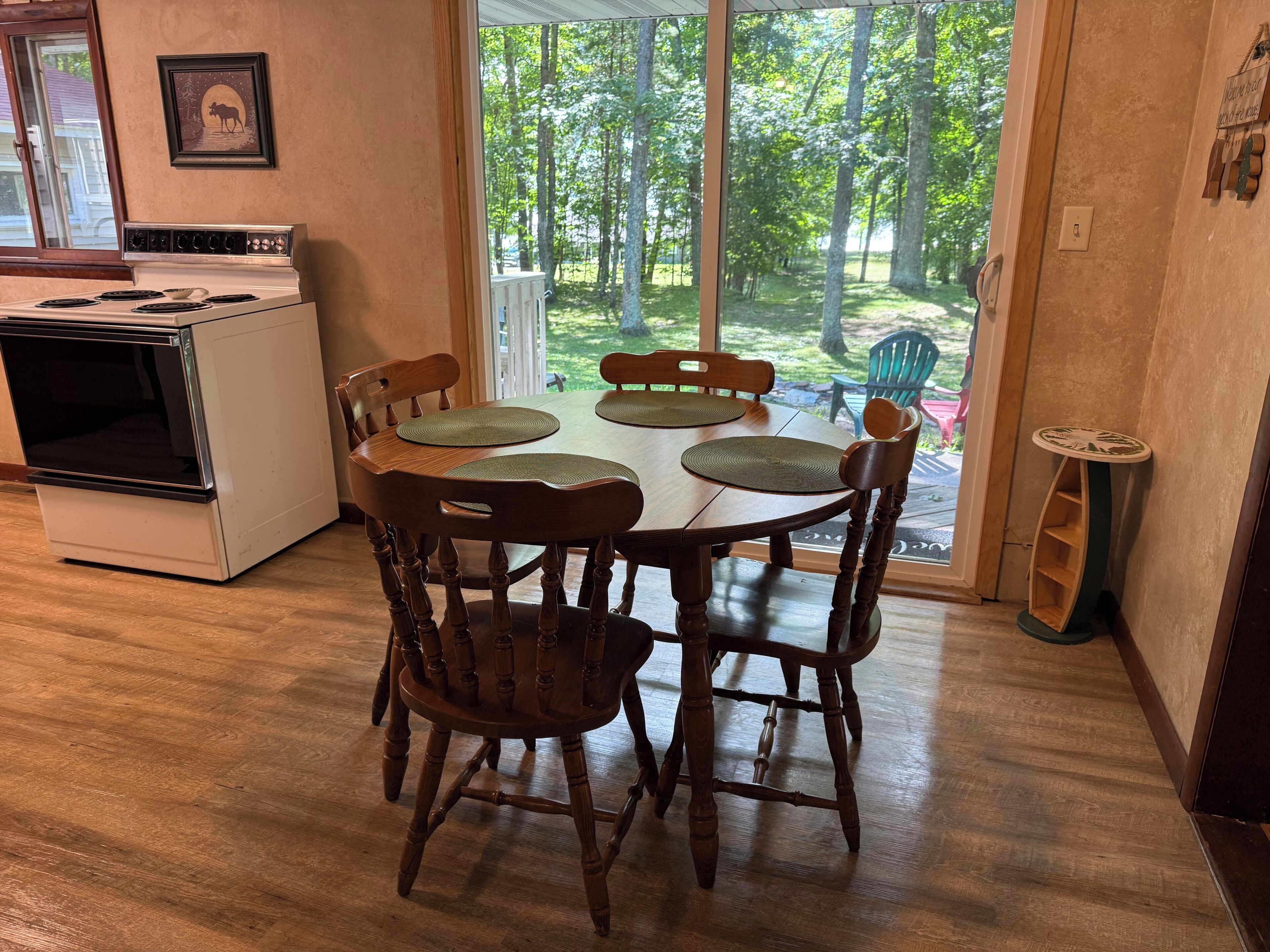 A dining area with a wooden table and chairs, a kitchen with a stove, and a view of a wooded area through a glass door.