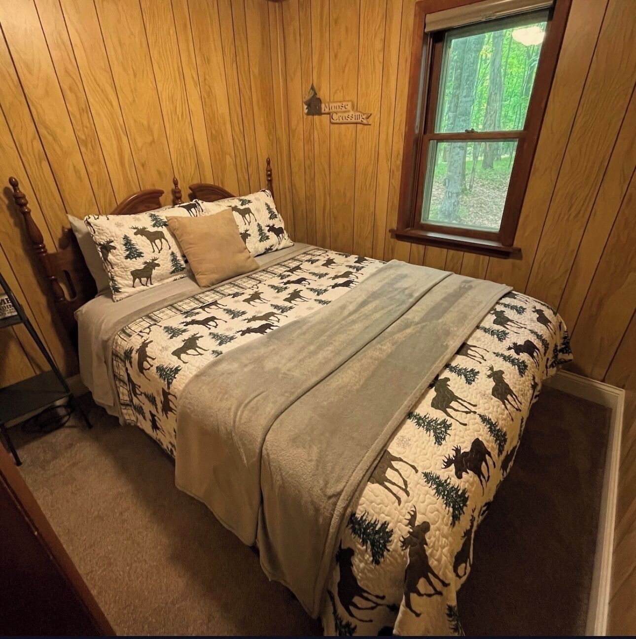 Una habitación con paneles de madera, una cama con colcha temática de alces, una ventana con vista a árboles y un letrero en la pared que dice "Moose Crossing".