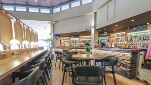 A spacious bar area with multiple seating options, including bar stools and round tables. There are various bottles and glasses on shelves, and a counter with a menu board. - 8 Puffin Way (Llwyngwril)