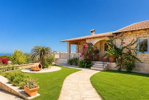 Una casa con fachada de piedra, techo de tejas, entrada pavimentada y un jardín bien cuidado con diversas plantas y flores.