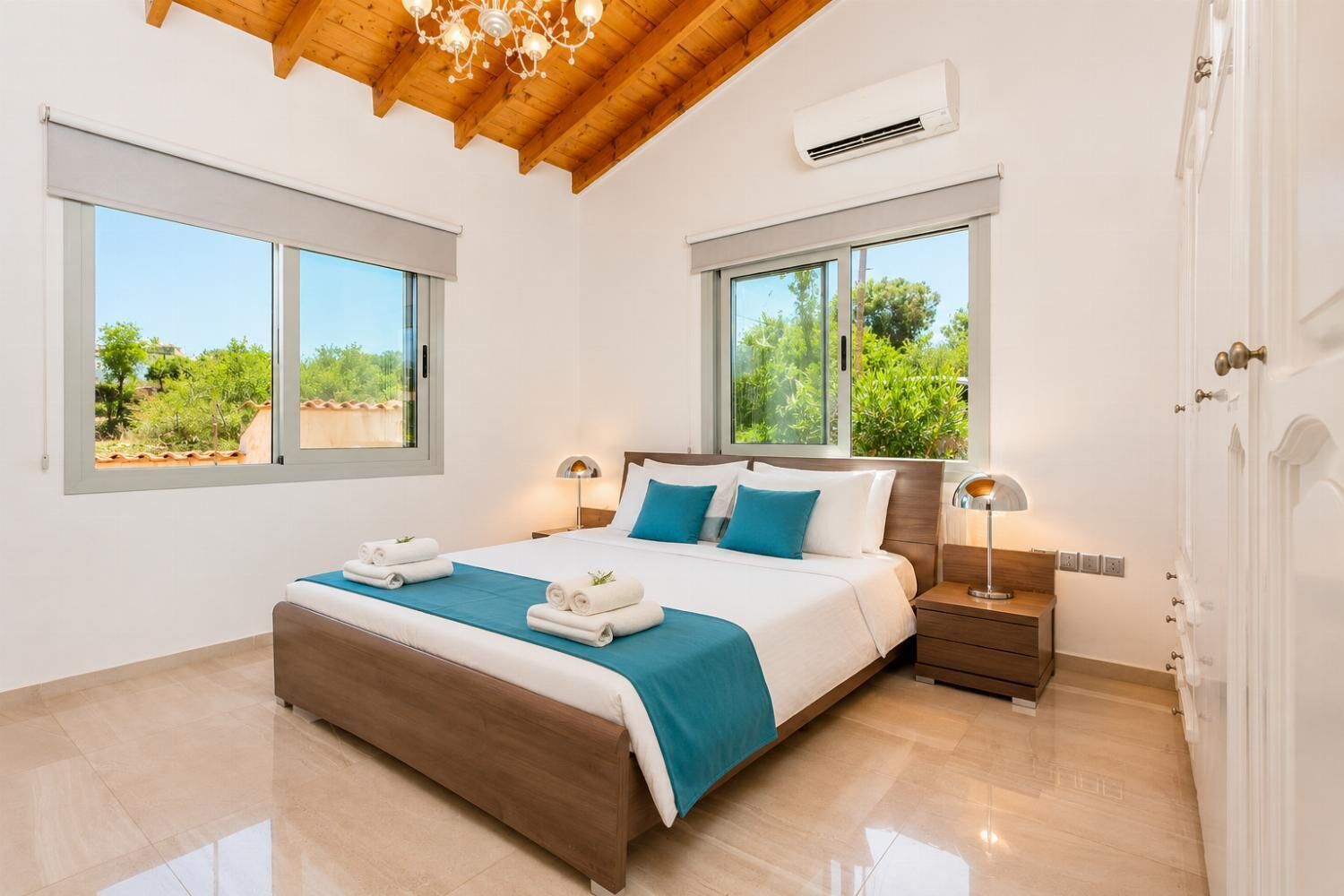 Une chambre avec un plafond en bois, deux fenêtres donnant sur la verdure, un lit avec une literie blanche et bleue, et une table de chevet avec une lampe.