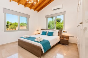 Un dormitorio con techo de madera, dos ventanas con vistas a la vegetación, una cama con ropa de cama blanca y azul, y una mesita de noche con una lámpara.
