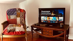 Een woonkamer met een televisie waarop een streamingdienst wordt weergegeven, een houten stoel met een kleurrijke deken en een houten bijzettafeltje met een afstandsbediening.