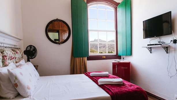 Quarto de hotel com cama, espelho, janela com persianas verdes, televisão e um armário vermelho.