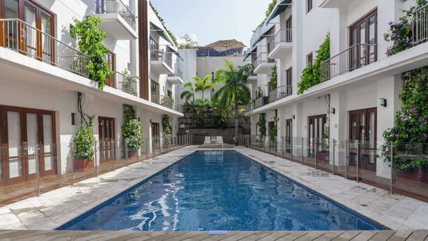 A swimming pool with a wooden deck, surrounded by white buildings with balconies and plants.