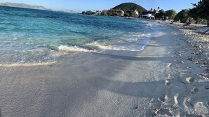 A beach with clear water, footprints, and buildings in the background.
